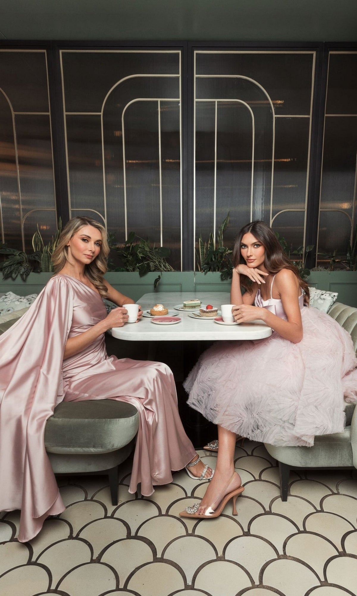 Two women wearing Short Homecoming Dress 20583 by Mac Duggal in rose pink sit at a round table in a chic café, enjoying coffee and pastries together.
